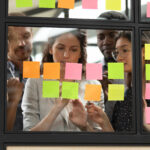 Focused young diverse teammates working with caucasian team leader on IT startup project kanban organization process, discussing details and tasks, writing notes on colorful paper stickers at office.